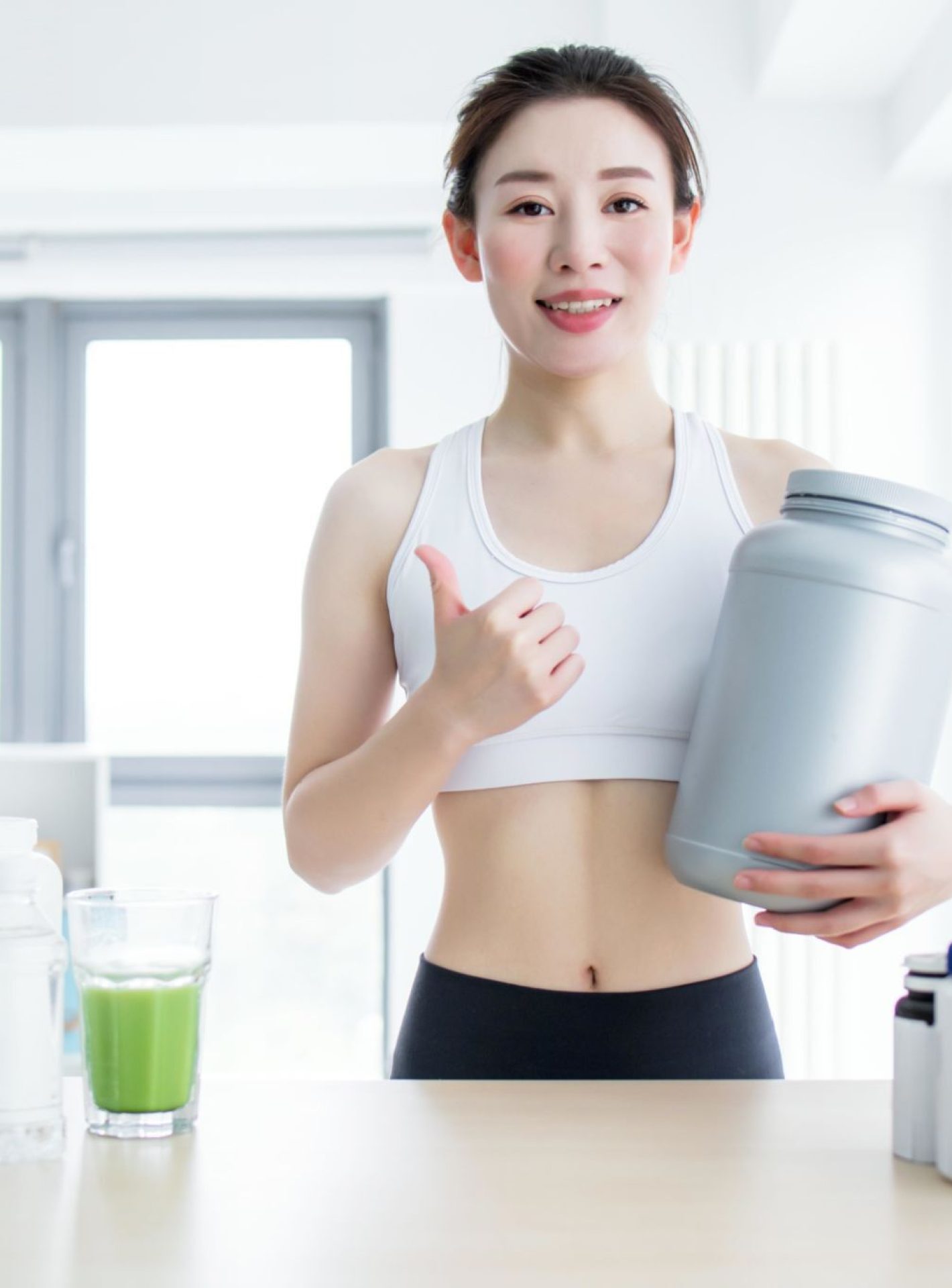 Asian woman holding a bottle of protein powder with various supplements on the table